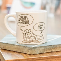 Caneca impressa com cena humorística de diálogo extraído do livro "Úrsula: uma peixinha do Sertão Carioca". A cena contém um diálogo entre a menina Maya e a peixinha Úrsula. Folheando um livro de biologia, Maya diz à Úrsula: "Aqui diz que você é um predador oportunista". E Úrsula responde: "Logo eu?!"