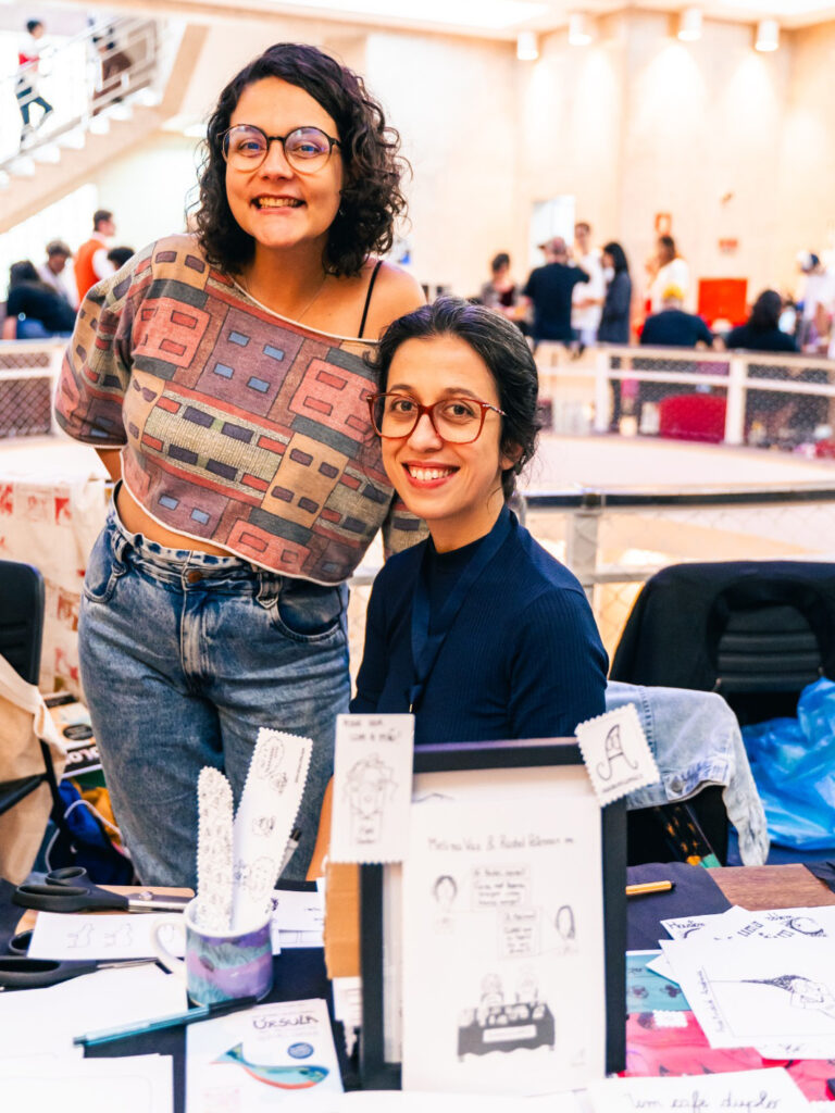 Melina Vaz (em pé e à esquerda) e Rachel Paterman (sentada e à direita) na banca Academicomics na Feira Miolos de 2024 (São Paulo)